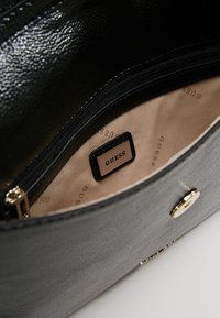 Grå faux läderhandväska med svart dragkedjeficka på insidan. Har "GUESS" logotag och beige foder med subtilt mönster av varumärket. Guldhardware-accent.