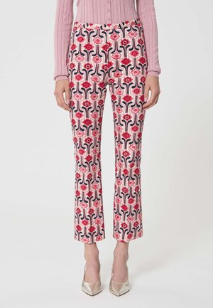 Femme portant un pantalon taille haute à motif floral rose et rouge, assorti d'un pull rose et de talons pointus métalliques, debout devant un fond blanc.