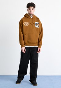 Sudadera con capucha marrón y bolsillo frontal, con texto blanco y un logotipo gráfico de la letra "C"; combinada con pantalones negros y zapatos negros.