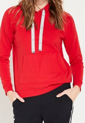 Woman wearing bright red hoodie with white text on drawstrings "CASUAL IDENTITY" and black pants with white stripes, hands in pockets.