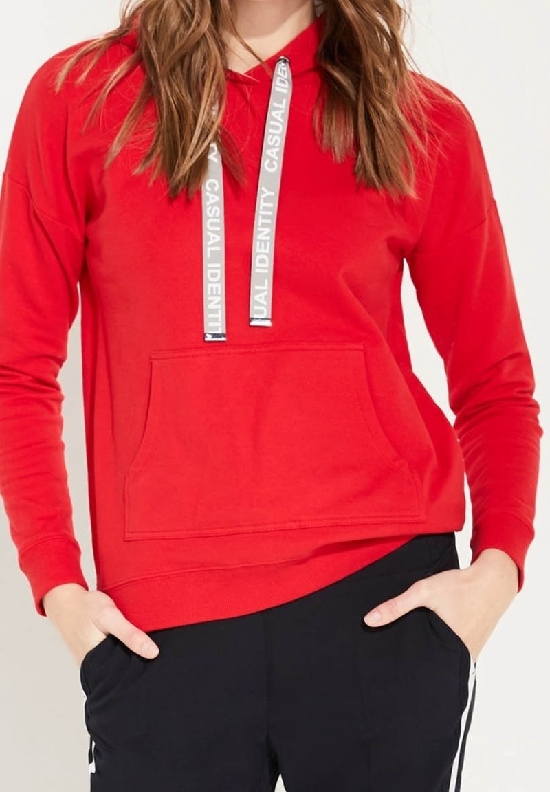 Femme portant un sweat à capuche rouge vif avec un texte blanc sur les cordons de serrage "CASUAL IDENTITY" et un pantalon noir à rayures blanches, mains dans les poches.
