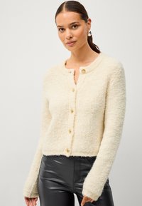 Cardigan tricoté crème à col rond, avec cinq boutons dorés et des manches longues. La texture est duveteuse et douce.