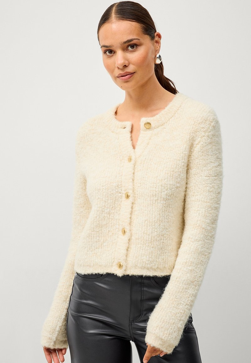 Cardigan tricoté crème à col rond, avec cinq boutons dorés et des manches longues. La texture est duveteuse et douce.