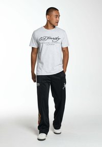 Camiseta de algodón gris con logotipo en negro, combinada con pantalones de chándal negros que presentan detalles bordados en rojo y blanco y zapatillas blancas.