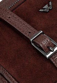 Bruin suède materiaal met een textuur leerensluiting, voorzien van een glanzende zilveren gesp en een embleemaccent, die rijke kleur en detail toont.