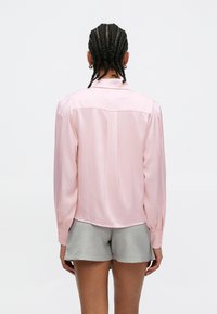 Camisa de cetim rosa claro com mangas compridas, corte relaxado e prega nas costas. Combinada com calções cinzentos, destacando textura suave e brilho subtil.