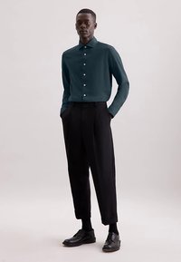 Donker teal overhemd met knopen en een spitse kraag, gecombineerd met zwarte, hoog getailleerde broek en zwarte leren schoenen, tegen een neutrale achtergrond.