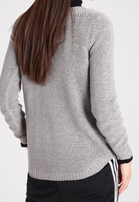 Szary dziergany sweter ze wzorem strukturalnym, z prążkowanymi mankietami i ściągaczem u dołu. Ma luźny krój i okrągły dekolt, stylizowany z czarnymi spodniami sportowymi.