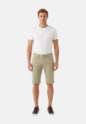 Uomo in piedi rivolto in avanti, indossa una maglietta bianca, pantaloncini beige al ginocchio e scarpe da ginnastica marrone scuro su uno sfondo semplice.