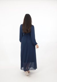 Vrouw die wegloopt, gekleed in een lange marineblauwe jurk met lange mouwen en transparante sandaaltjes met hak, tegen een witte achtergrond.