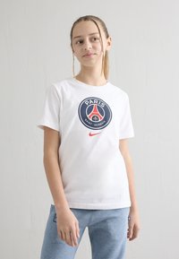 Tienermeisje met gevlochten haar draagt een wit Nike Paris Saint-Germain T-shirt en lichtblauwe joggingbroek, staand tegen een witte muur.