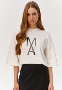 Biały skrócony t-shirt z czarnym graficznym wzorem "MA", szerokie rękawy, materiał bawełniany, noszony na czarnej spódnicy, gładka tekstura.
