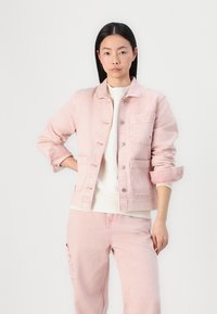 Chaqueta de mezclilla rosa con cierre de botones, dos bolsillos frontales, mangas dobladas y un corte ligeramente holgado. Llevada sobre una camiseta blanca.