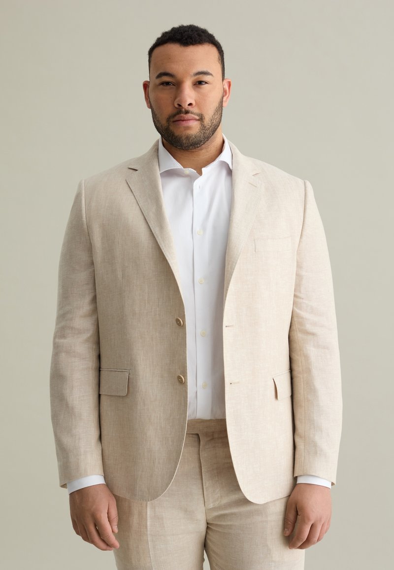 Costume en lin beige avec une veste à simple boutonnage et un pantalon assorti. Comprend deux boutons, des revers crantés et une chemise blanche à col standard.
