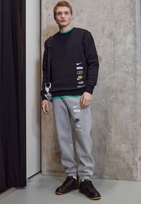 Felpa nera con loghi Nike stampati, colletto interno verde, pantaloni grigi con branding simile e scarpe nere con suole di gomma.