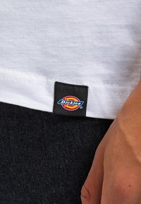 Hvid t-shirt med synlige søm i kanten og et sort stofmærke med "Dickies" logo i farvet cirkeldesign.