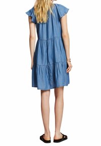 Robe en denim avec des volants superposés, des manches courtes et flottantes, et une coupe décontractée. Couleur bleu clair avec une texture douce. Portée avec des sandales noires.