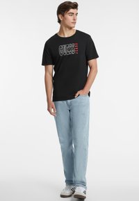 Juodas medvilnės marškinėliai su balta ir raudona "GUESS JEANS" logo, derinami su šviesiai mėlynomis džinsinėmis kelnėmis ir baltais sportbačiais.