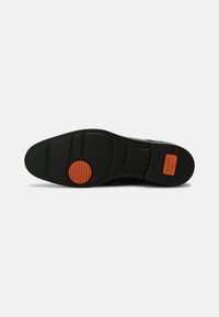 Musta kumipohja, jossa on tekstuuriset kuviot ja keskellä pyöreä oranssi aksentti, jossa on brändilogo kulmassa.