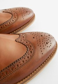Zapatos de vestir de cuero marrón con detalles perforados, punta afilada y un acento cosido a lo largo de la suela para mayor textura.