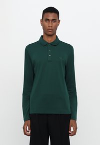 Grønn langermet polo skjorte laget av myk stoff, med krage, plakk med tre knapper og en liten logo på venstre bryst.