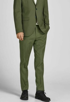 Homme portant un costume vert olive avec des chaussures noires, debout avec une main dans la poche devant un fond gris.