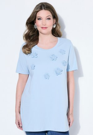 Frau trägt ein hellblaues Kurzarmshirt mit erhabenen Blumenapplikationen, kombiniert mit blauen Jeans, vor weißem Hintergrund.