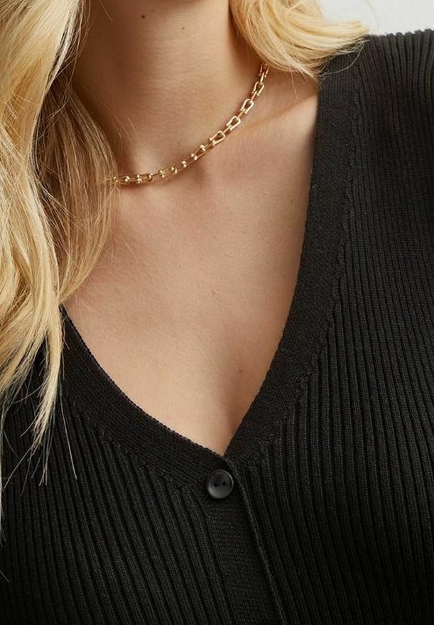 Collier chaîne en or porté par une personne vêtue d'un haut noir côtelé à col en V boutonné, avec de longs cheveux blonds tombant sur une épaule.
