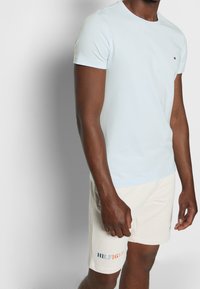 Hellsblaues Baumwoll-T-Shirt mit Rundhalsausschnitt, kombiniert mit cremefarbenen Shorts, die ein farbenfrohes "HILFIGER"-Logo am linken Bein aufweisen.