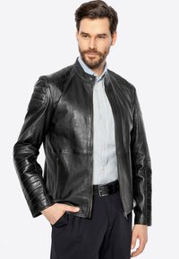 Giacca in pelle nera con colletto alto, zip frontale, dettagli a spalline trapuntate e due tasche laterali. Texture liscia e design aderente.