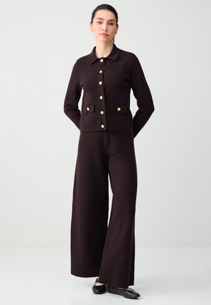 Donna che indossa un cardigan marrone scuro con bottoni dorati, pantaloni ampi coordinati e scarpe nere piatte, in piedi davanti a uno sfondo neutro.