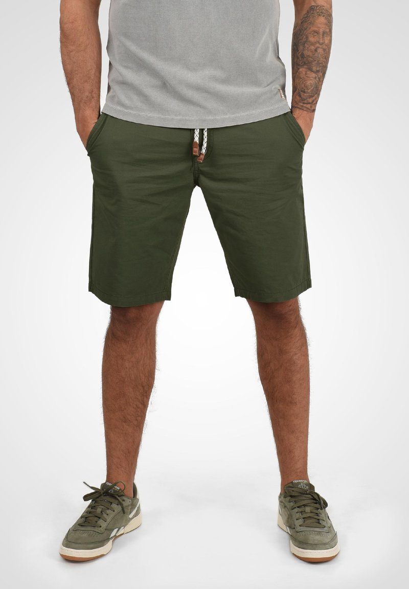 Bărbat purtând pantaloni scurți verde măsliniu cu șnur alb, tricou gri, tatuaje pe brațul drept și adidași verzi, stând cu mâinile în buzunare.