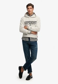 Lichtgrijze hoodie met het "SUPERDRY"-logo, gedragen over een geruit overhemd. Donkerblauwe spijkerbroek en zwarte loafers maken de outfit compleet.