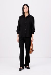 Black button-up shirt with a relaxed fit and rolled sleeves, paired with black pants and black studded flats. Brown shoulder bag as an accent.