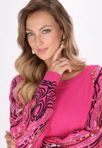 Pullover rosa con maniche lunghe caratterizzato da un design a fantasia in nero e paillettes metalliche dorate e argentate. Modella in posa con la mano sul mento.