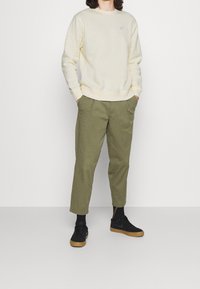 Sudadera amarilla claro con cuello redondo y pequeño logo, combinada con pantalones verde olivo ajustados y zapatillas negras con suelas de goma.