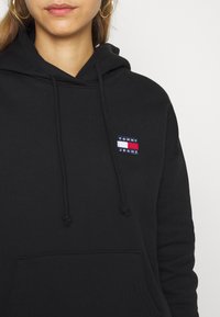 Svart hoodie med dragsnören, med ett litet broderat märke "TOMMY JEANS" i marinblått, vitt och rött. Materialet verkar mjukt och strukturerat.