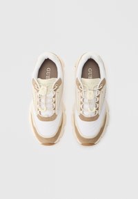 Μπεζ και λευκά αθλητικά παπούτσια Guess με χοντρούς πάτους και ελαστικά κορδόνια χωρίς δέσιμο, σε άποψη από πάνω σε απλό λευκό φόντο.