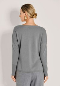 Frau mit blonden Haaren zu einem Dutt gebunden, die goldene Ohrringe, einen grauen Pullover mit langen Ärmeln und graue Hosen trägt, mit dem Rücken zur Kamera.