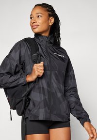 Jachetă ușoară de culoare neagră cu un model geometric, cu fermoar complet, guler înalt și detalii cu logo. Asortată cu un rucsac negru și șorturi.