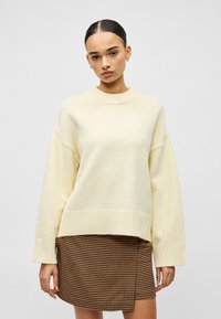 Κρεμ oversized πουλόβερ με ριμπ πλέξη στον λαιμό και τις μανσέτες, συνδυασμένο με μια καφέ καρό φούστα με σχέδιο τύπου wrap.