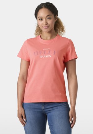 Kobieta o kręconych włosach, ubrana w koralowy T-shirt Helly Hansen i niebieskie dżinsy, stoi na jednolitym jasnym tle.