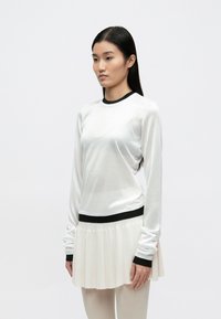 Balts garām piedurknēm tops ar melnu apkakli un aprocēm, kombinēts ar krokotu krēmkrāsas svārku. Audums šķiet viegls ar gludu tekstūru.