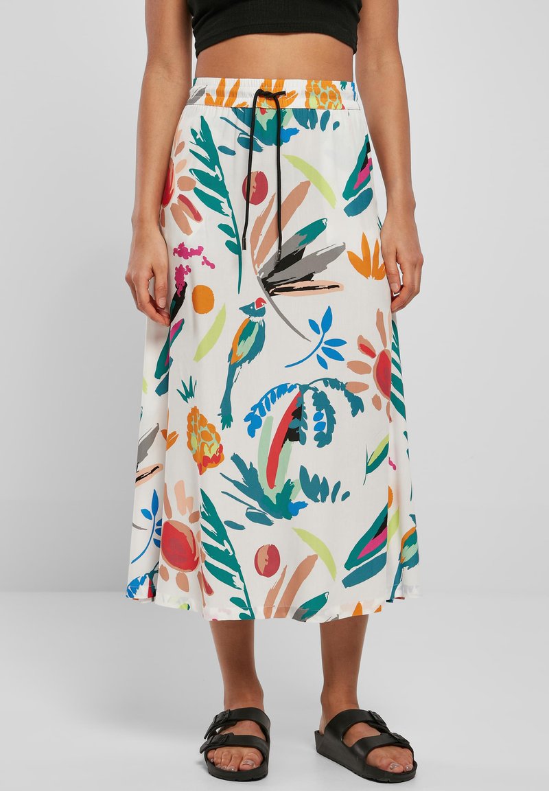 Mujer con falda midi blanca con patrones tropicales coloridos y cordón negro, combinada con sandalias negras y un top corto negro.