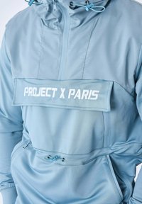 Ljusblå vindjacka med hög krage, dragskohette, framficka och vit "PROJECT X PARIS" textpatch. Slät, vattentålig tyg.