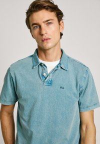 Svetlomodrá polo košeľa vyrobená z látky podobnej denimu. Má golier, krátke rukávy a jemne vyšívané logo na hrudi.