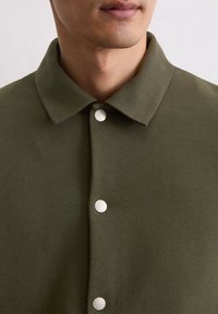 Zielona oliwkowa koszula z kołnierzykiem, z metalowymi zapięciami i gładkim, teksturowanym materiałem. Zbliżenie na górną część odzieży.