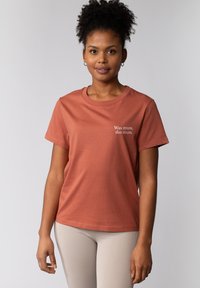 T-shirt de couleur rouille en coton, avec des manches courtes et un col rond. Il y a un texte blanc à l'avant : "Was muss, das muss."