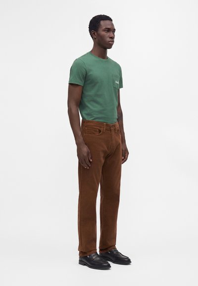 Uomo in piedi che indossa una camicia verde a maniche corte, pantaloni marroni e scarpe nere su uno sfondo chiaro uniforme.