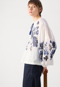 Weiße Baumwollbluse mit blauen Blumenstickereien, mit Puffärmeln, Bindungsdetail mit Quaste, kombiniert mit dunklen Jeans.